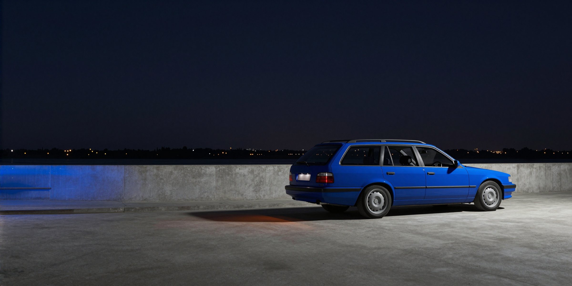 A deep blue estate car seen from behind at night beside a low concrete parapet, the lights of a distant city low along the horizon