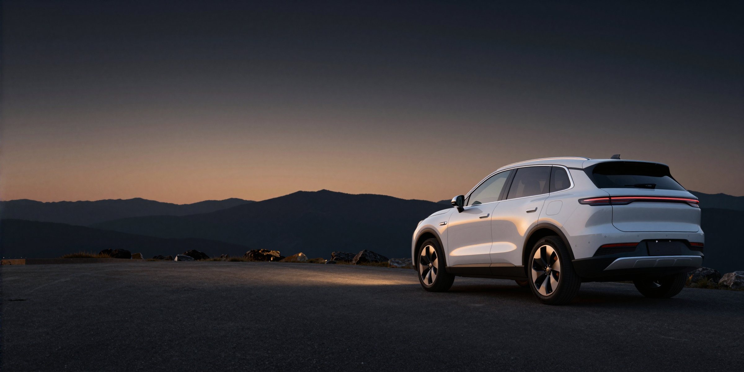 A pearl white SUV seen from behind on a mountain road at dusk, the sky fading to amber over distant ridges