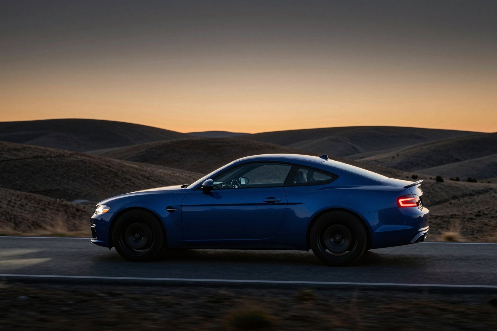 A deep blue coupé driving along an empty road through rolling hills at dusk, its tail lights glowing
