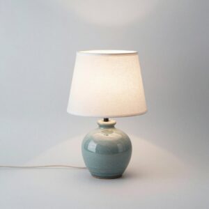 Table lamp with a rounded pale sea-green stoneware base and an off-white linen drum shade, switched on