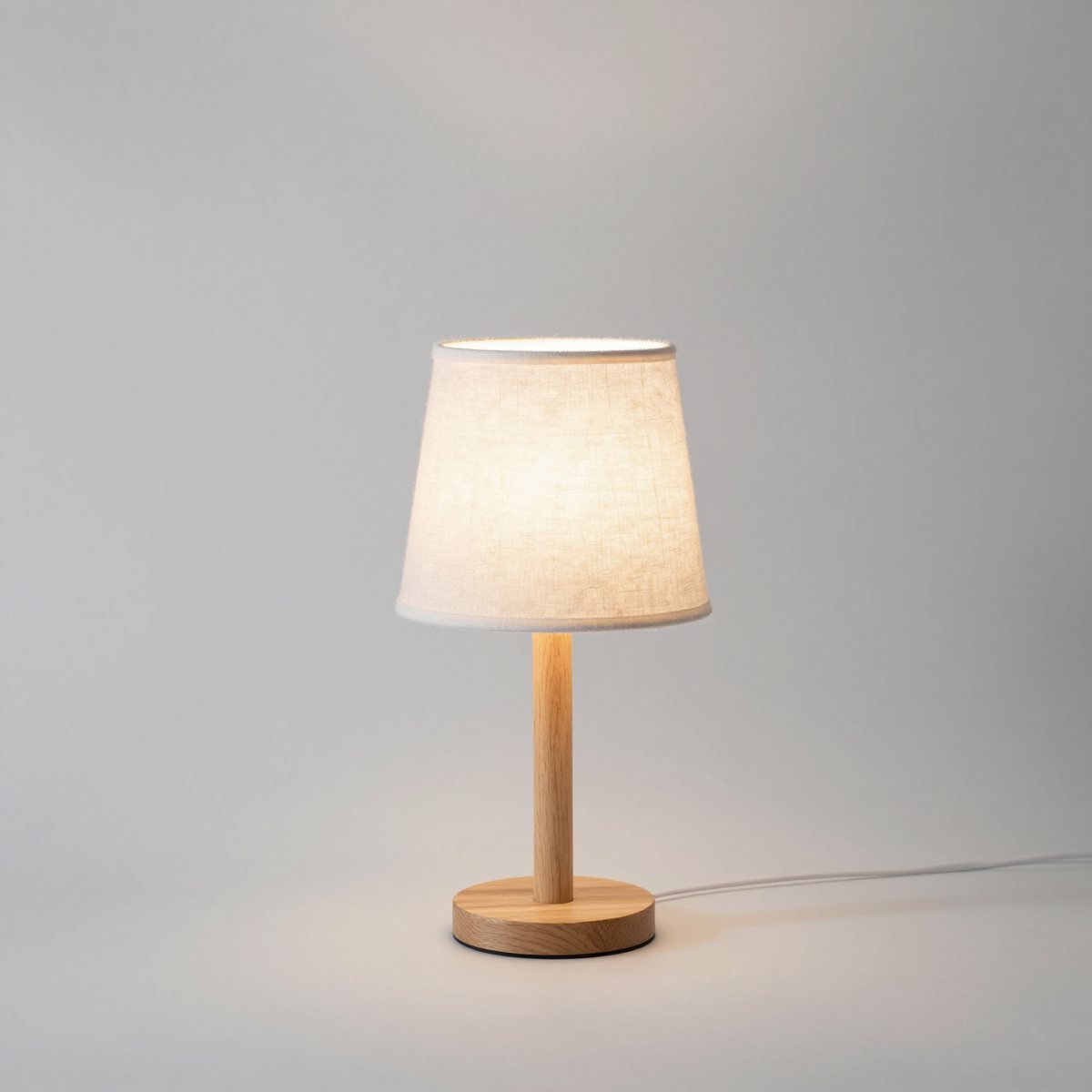 Small oak bedside lamp with a straight stem, a round base and a linen drum shade, switched on