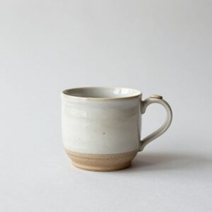 Speckled stoneware mug in a creamy off-white glaze over a bare clay foot, with a thumb rest on the handle