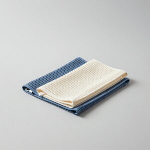 Two waffle-weave tea towels, one denim blue and one natural, folded and stacked, a hanging loop showing