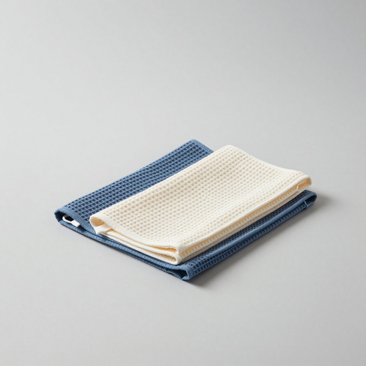 Two waffle-weave tea towels, one denim blue and one natural, folded and stacked, a hanging loop showing